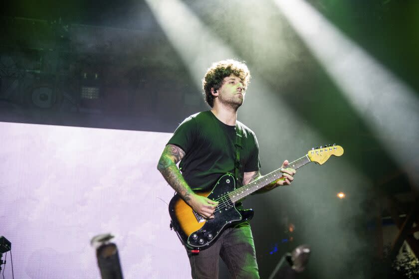 A man with short brown hair wearing a black T-shirt and jeans while playing guitar on a stage