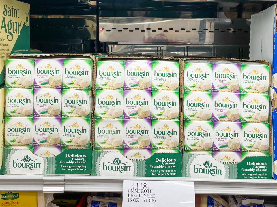 Three-packs of Boursin cheese, two of which are green and one with purple packaging, in a larger green cardboard box on shelf at Costco