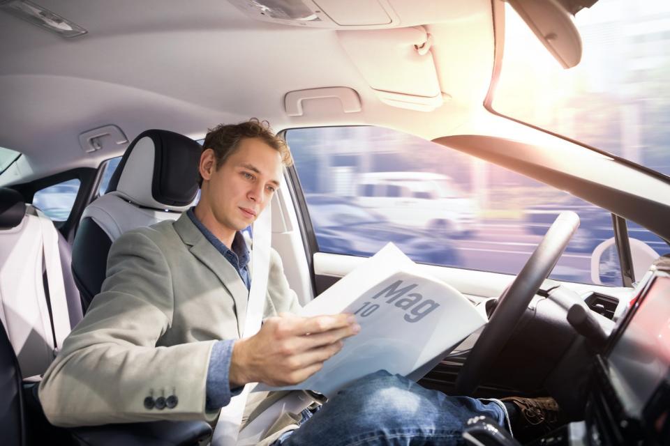 A driver reading paperwork behind the wheel of an autonomous vehicle driving itself down a road. 