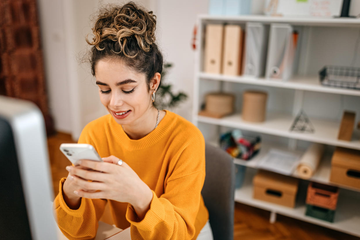 Confident young female entrepreneur wearing yellow sweatshirt using smart phone while sitting by computer at desk in office