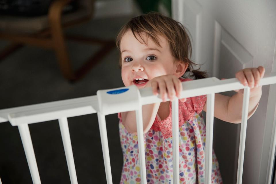 Front view of a little girl standing on the other side of a baby saftey gate. The little girl is holding onto the top of the saftey gate and looking at the camera over the top.