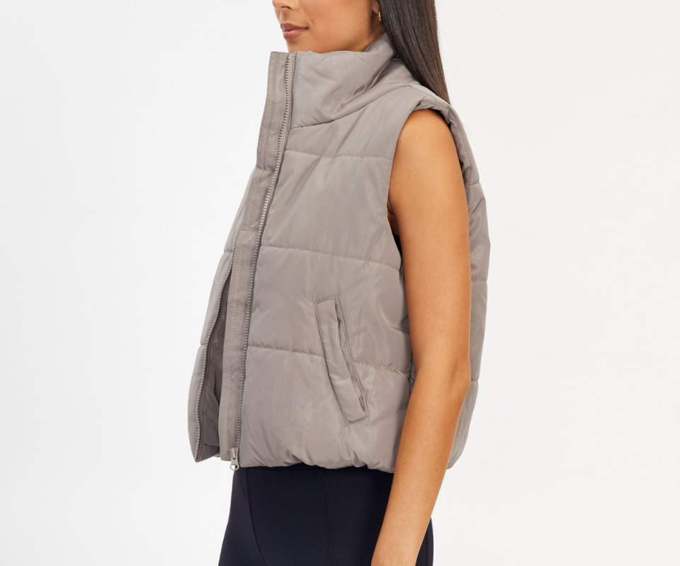 An olive skinned girl stands to her side with a smokey grey puffer vest against a white background.