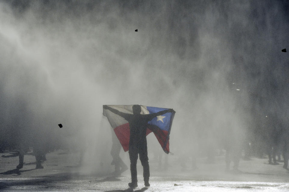 FILE - In this Oct. 25, 2019 file photo, an anti-government protester holds out a Chilean flag during clashes with police in Valparaiso, Chile. Student protests over a fare hike morphed into a nationwide call for socio-economic equality and better social services that brought millions to the streets and forced President Sebastián Piñera to increase benefits for the poor and disadvantaged and start a process of constitutional reform. (AP Photo/Matias Delacroix, File)