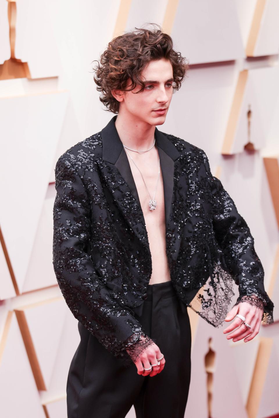Timothee in a sequin bedazzled cropped suit jacket with black lace on the arm cuffs and around the waist of the jacket. He has no shirt on and two silver necklaces. He's wearing black trousers.