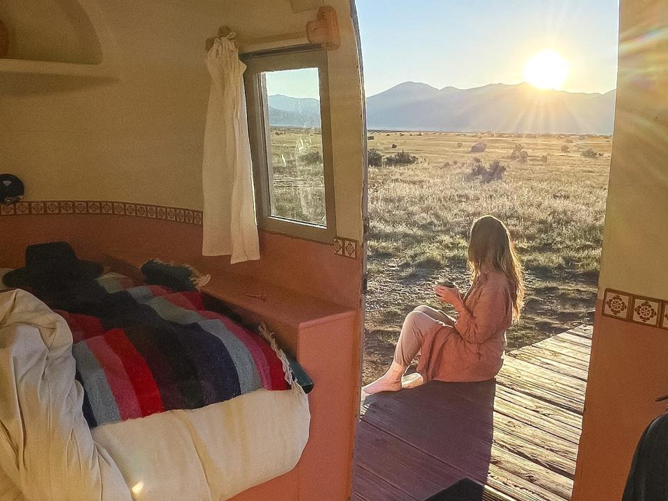 In the foreground is a small hotel room with an open door. Outside the door, Emily sits on a wooden path with a cup of coffee.  She is looking out at a wide-open area with mountains.