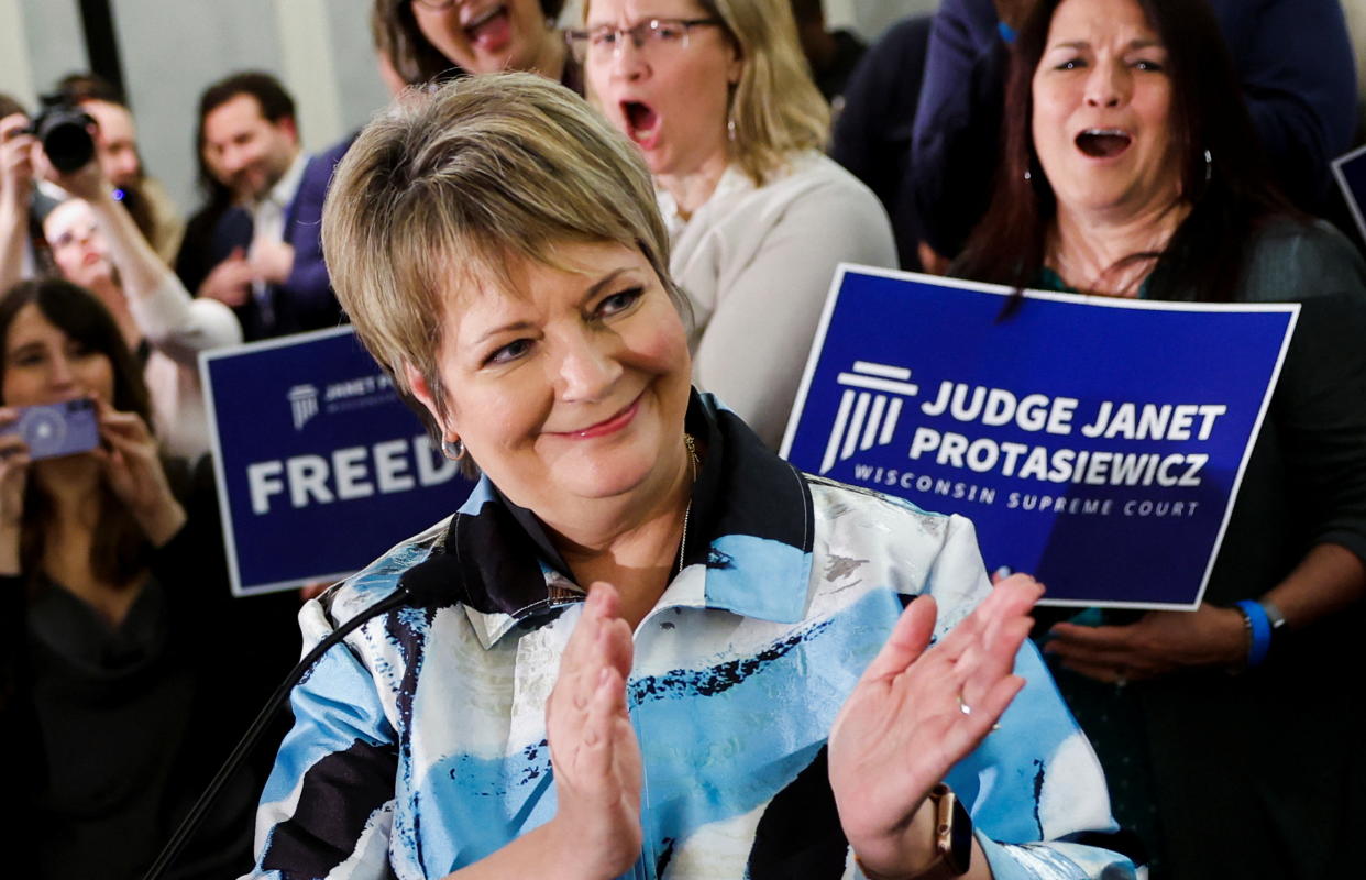 Janet Protasiewicz, wearing a broad smile, claps as her supporters cheer her on in the background.