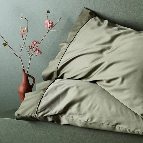 A pair of jade colored pillowcases in a soft silk sitting next to sprigs of cherry blossom in a funky vase