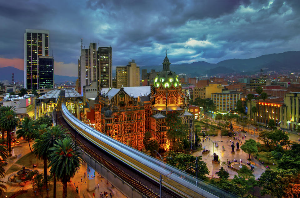 The elevated Medellin Metro is in motion as it rushes into the Parque Berrio Station in front of the illuminated Palace of Culture in Plaza Botero in Medellin, Colombia.  The City of Eternal Spring is located in the Aburra Valley, a central region of the Andes Mountains in South America.