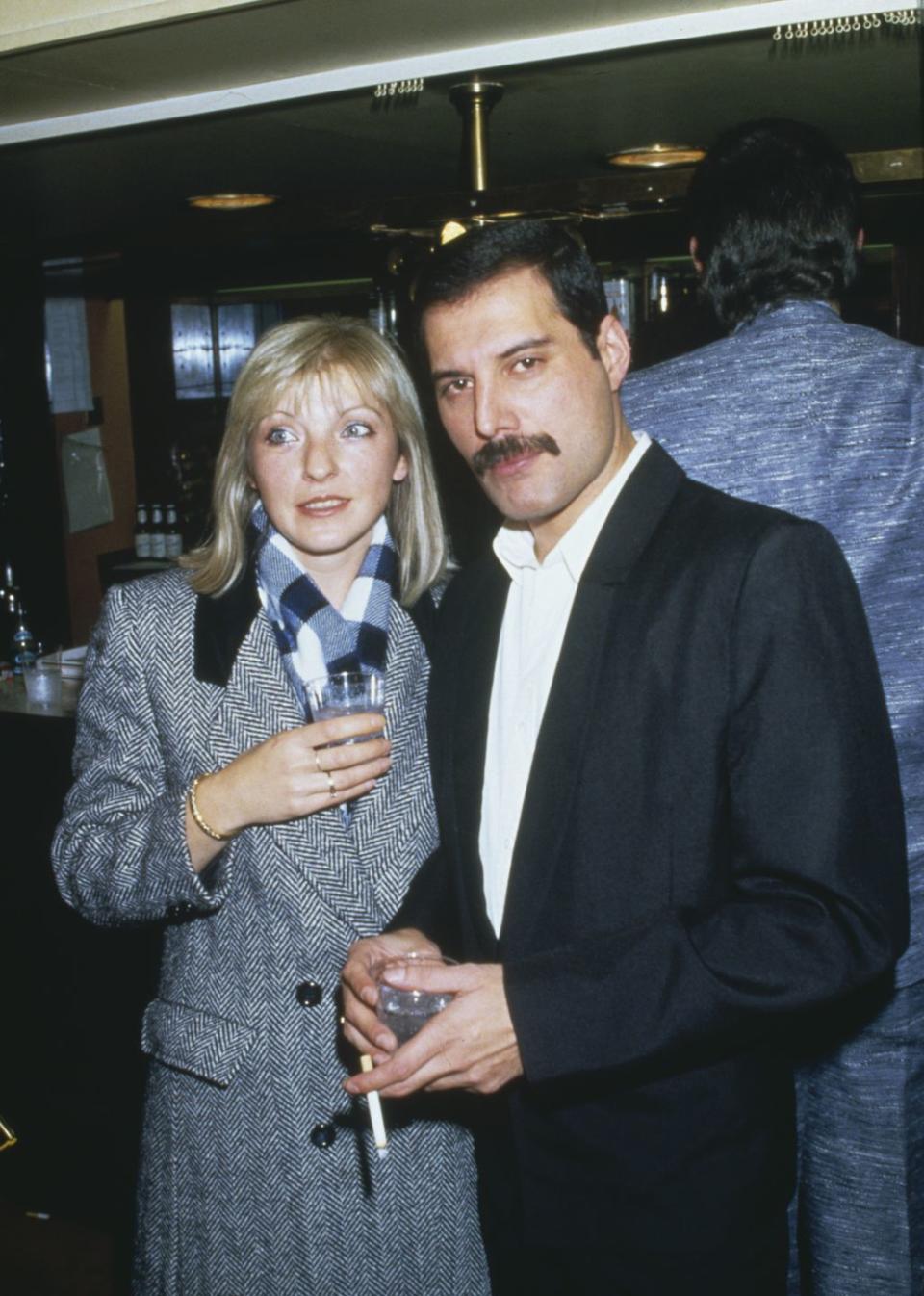 mary austin and freddie mercury stand next to each other at an event, both are holding drinks and mercury has a cigarette in one hand, she is wearing a chevron black and white jacket with a plaid scarf, he is wearing a black suit without a tie