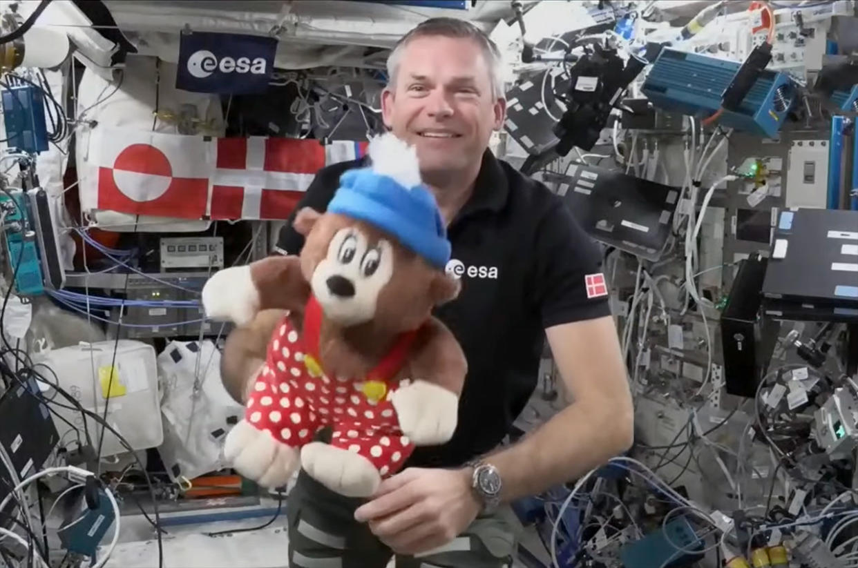  A man in a black polo shirt holds a plush bear inside the international space station. 