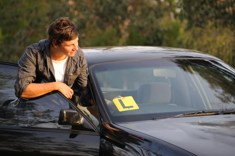 A young man looks stands outside his car with the front door open. A learner plate is on the windshield.