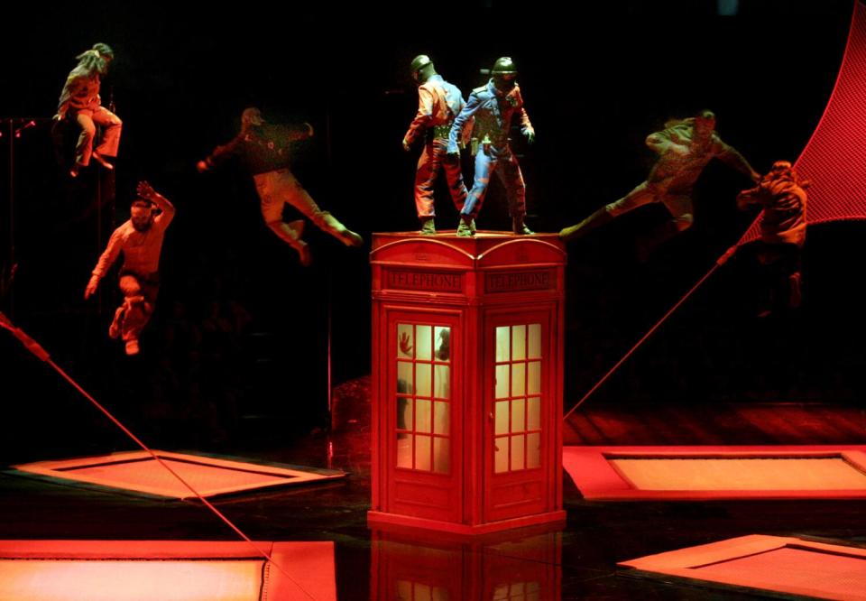 Performers stand on a red phone booth as others jump in the air.