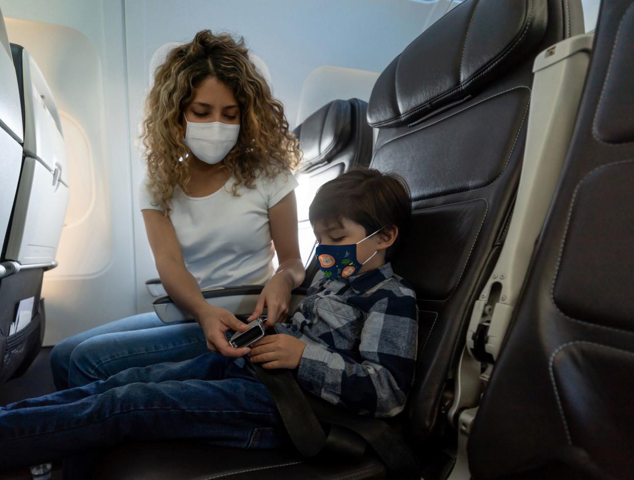 Latin American mother traveling by plane with her son during the pandemic and fastening his seat belt while wearing facemasks