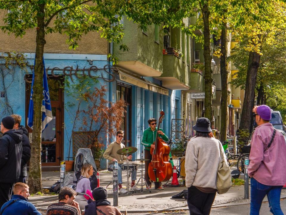 Musicians play on a lush street corner with pedestrians sitting and walking by.