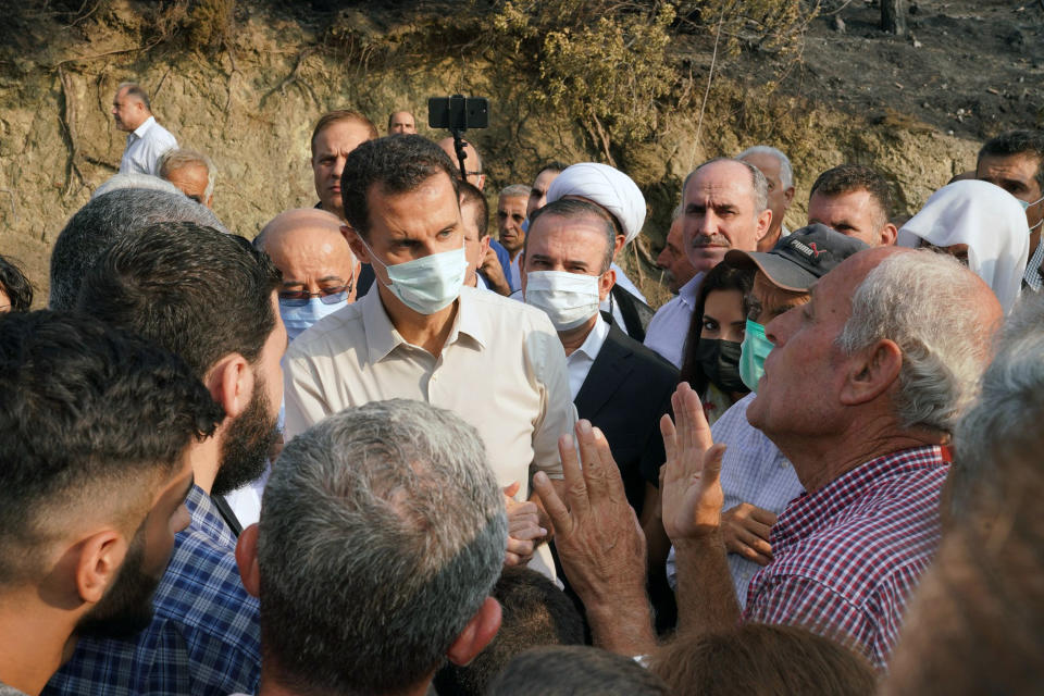 In this photo released Tuesday, Oct. 13, 2020 on the official Facebook page of the Syrian Presidency, Syrian President Bashar Assad, center, wearing a mask to help prevent the spread of the coronavirus, speaks with people during his visit to the coastal province of Latakia, Syria. Assad made a rare public visit to Latakia where he toured areas that suffered heavy damage in last week’s deadly wildfires. (Syrian Presidency via Facebook via AP)