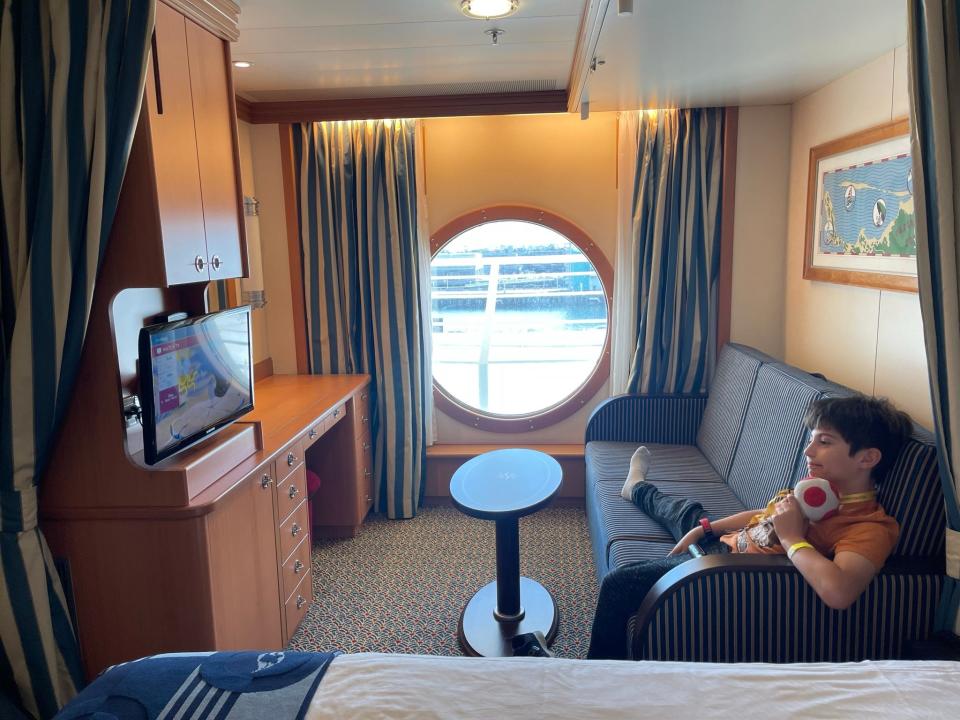 disney wonder interior stateroom living area with person sitting on couch, porthole window on back wall looking out to ocean