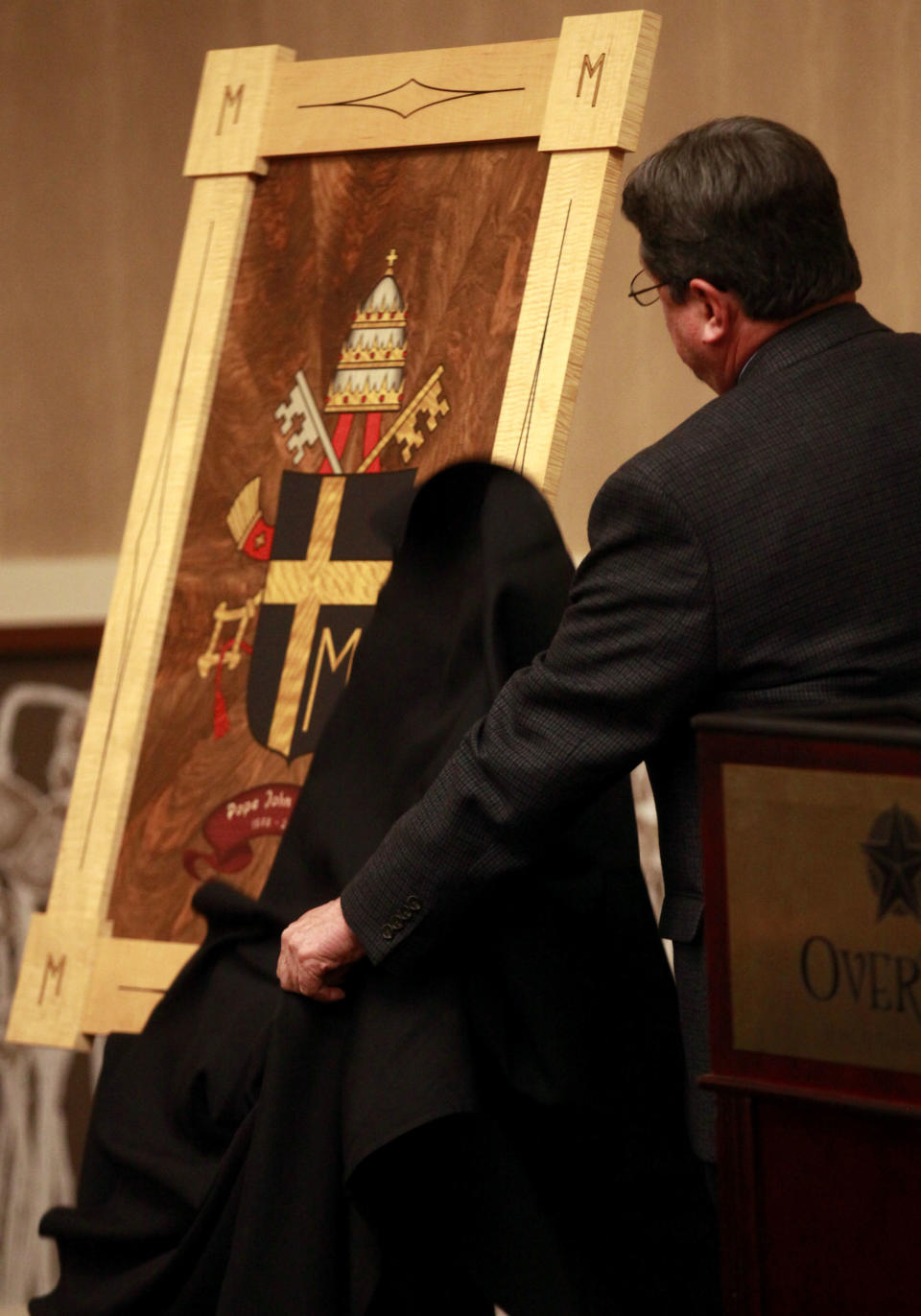 Chancellor of The Catholic Diocese of Lubbock Marty Martin unveils the coat of arms of Pope John Paul II at the Overton Hotel and Conference Center during an announcement of an exhibit displaying artifacts from Pope John Paul the II is coming to Lubbock, Thursday, Nov. 29, 2012. The coat of arms is inlay wood made from 21 different types of wood. Over 130 items are expected to be on display between March 15, 2013 and May 31, 2013 at The Catholic Renewal Center of the Diocese of Lubbock. (AP Photo/Lubbock Avalanche-Journal, Stephen Spillman)