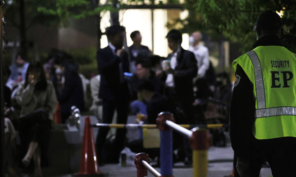 A security person stands guard as people drink at a park in Tokyo on April 21, 2021. Trains packed with commuters returning to work after a weeklong national holiday. Frustrated young people drinking in the streets because bars are closed. Protests planned over a possible visit by the Olympics chief. As the coronavirus spreads in Japan ahead of the Tokyo Olympics starting in 11 weeks, one of the world’s least vaccinated nations is showing signs of strain, both societal and political. (Takuto Kaneko/Kyodo News via AP)