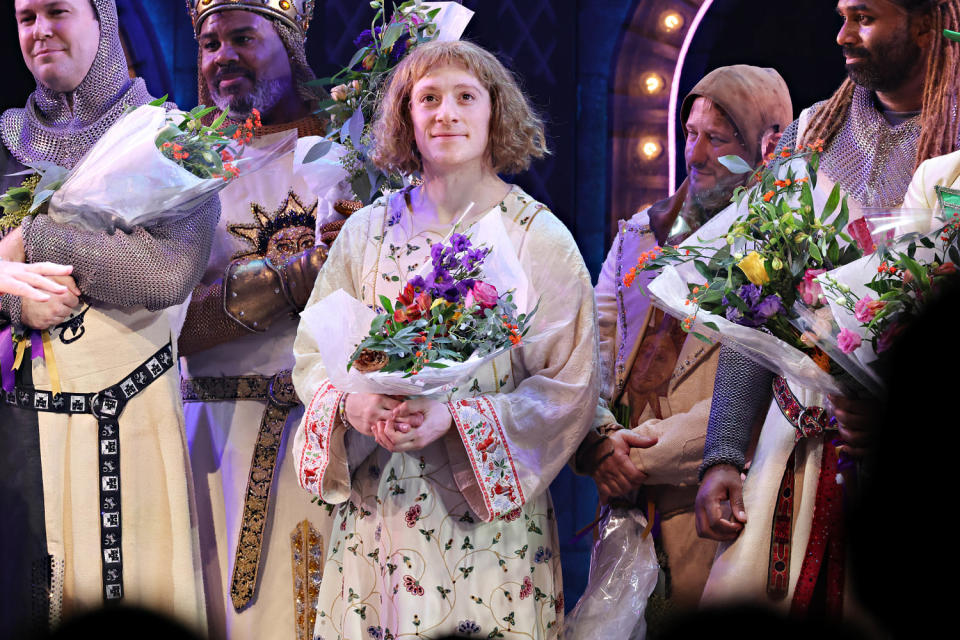 Ethan Slater holds flowers in an old-fashioned floral tunic costume and a bowl-cut wig. He's surrounded by other men in medieval looking knight's outfits also holding flowers. (Cindy Ord / Getty Images)