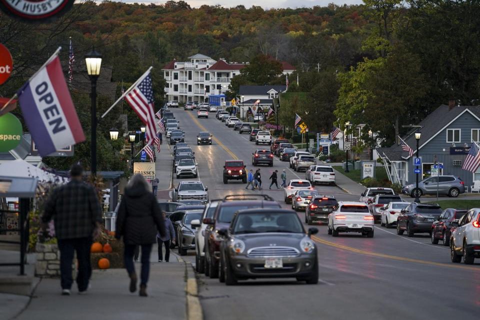 People walk down a street with stores on either side of it, with trees and a white building in the distance.