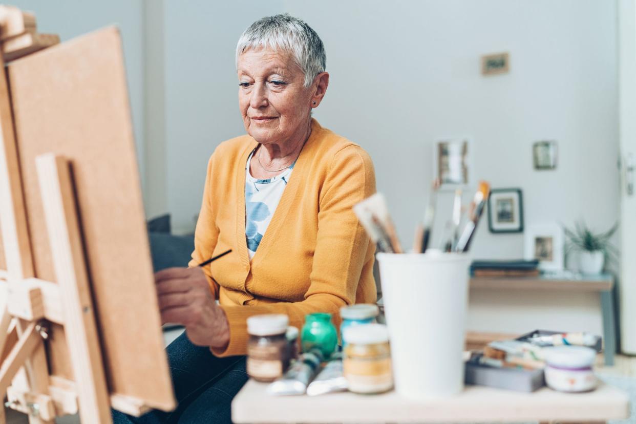 Focus on senior woman painting with back of easel holding the painting and table blurred in the foreground, holding paints and other paraphernalia for painting, wall and hanging pictures blurred in the background