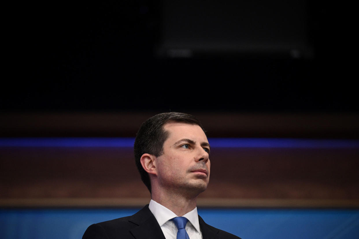 Transportation Secretary Pete Buttigieg in a suit with a white shirt and blue tie looks off to the side while delivering remarks during a press conference