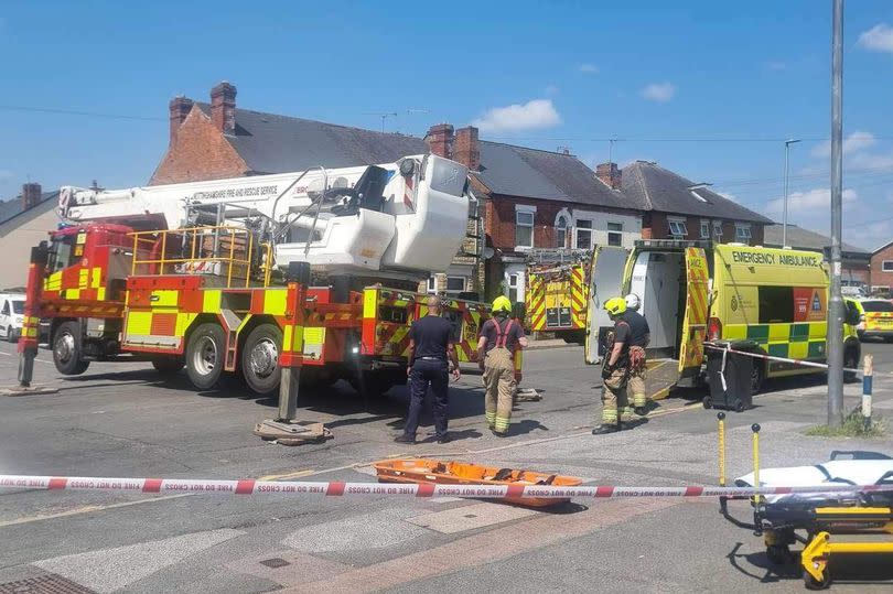 Fire engine with aerial ladder on top on left, ambulance with doors open on right, back of another fire engine visible behind and police car visible on right hand side, with multiple firefighters and other people including members of public visible and police tape cordoning off road with townhouses visible