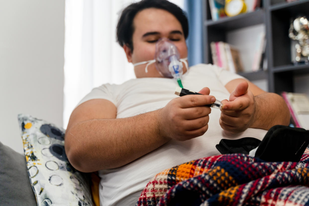young man sits in a sofa with an oxygen mask and  measuring blood sugar level