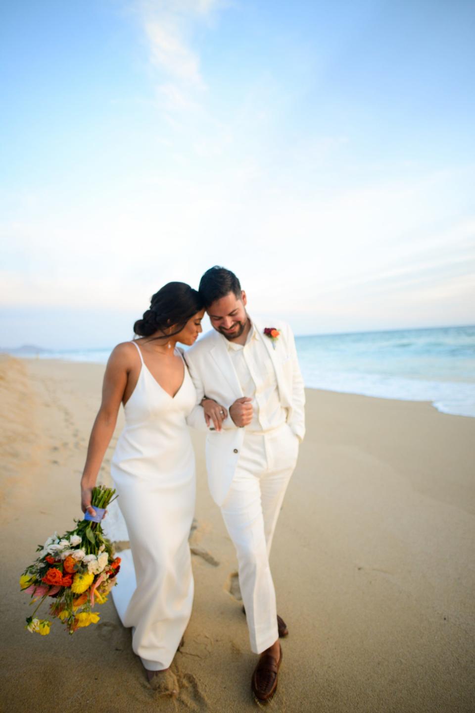 A man and woman lean their heads together as they walk with linked arms on a beach.