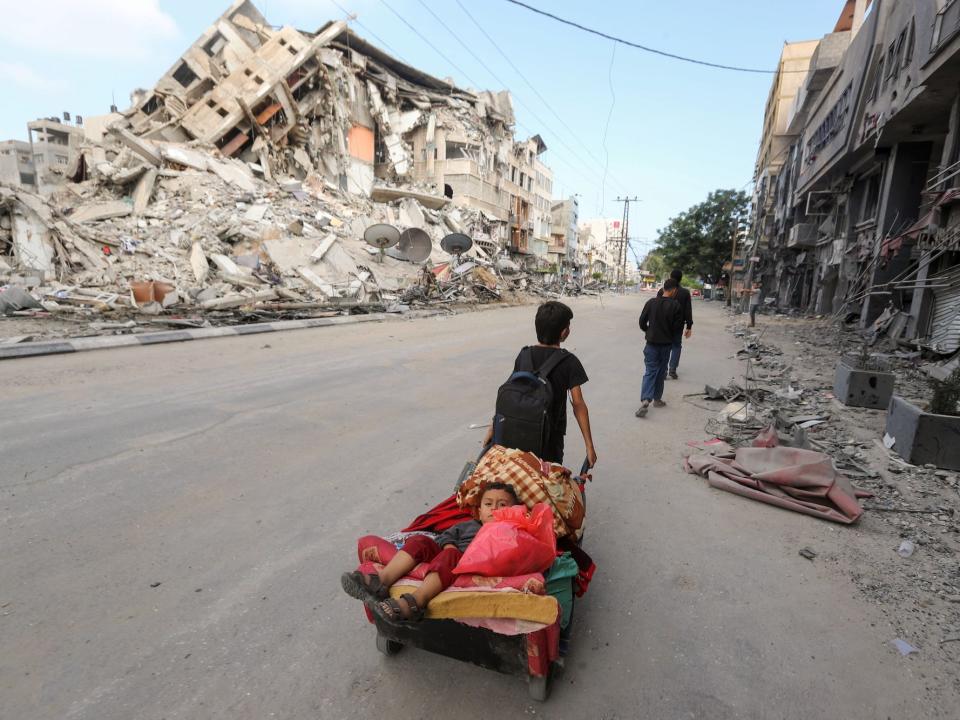 A Palestinian boy drags another boy on a cart in the street, with a ruined building in the background.