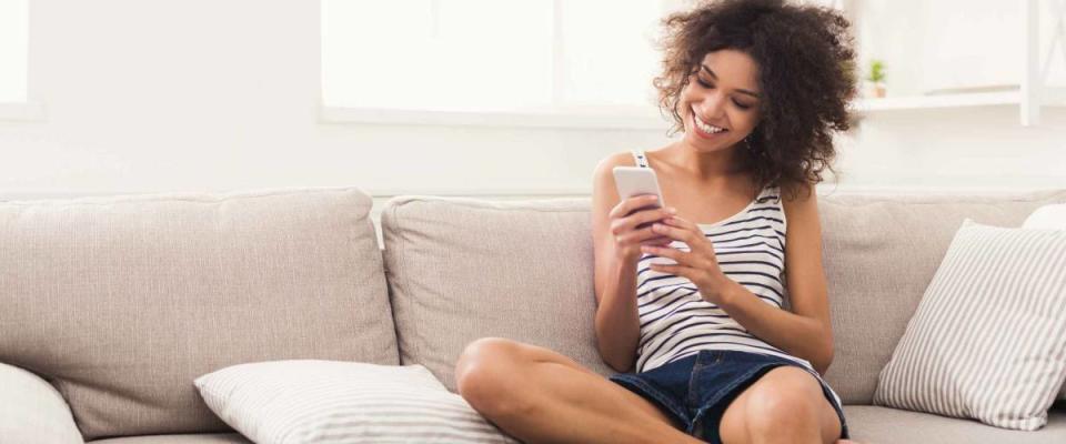 Social networks. Young black woman messaging on smartphone at home, sitting comfortably on beige sofa, copy space