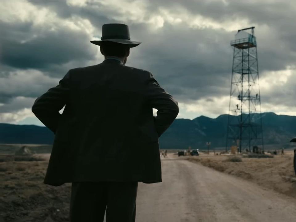 Cillian Murphy in a scene from "Oppenheimer" where J. Robert Oppenheimer is standing by the side of a dirt road next to a black car while gazing into the distance.