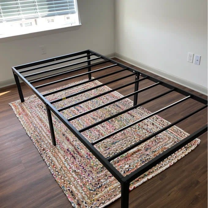 Reviewer's photo of the black metal bed frame on a colorful rug in a room with hardwood floors and blinds on window