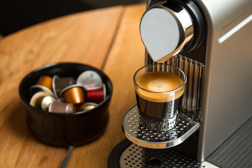 Freshly brewed espresso in glass standing on silver Nespresso coffee machine. Blurred background with Nespresso capsules in black bowl.