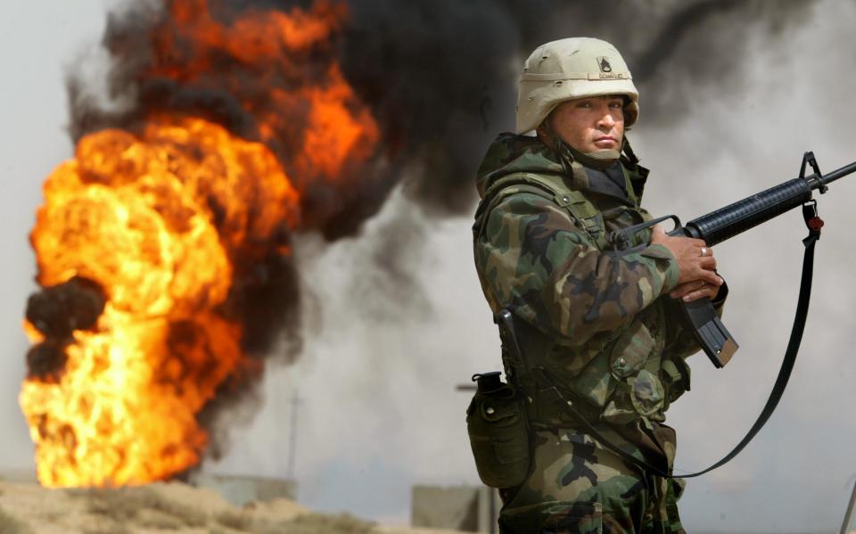 Sgt. Robert Dominguez, a massive blaze behind him, stands with his rifle at the ready and looks at the photographer.