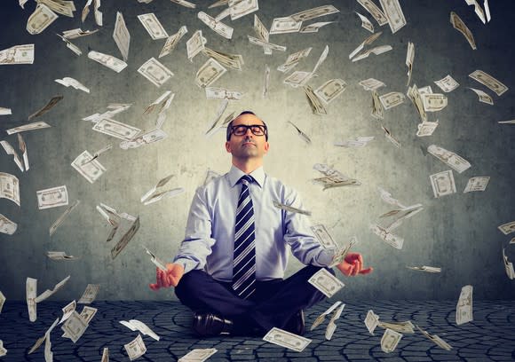 A man sits in a yoga pose as dollar bills fall down around him.