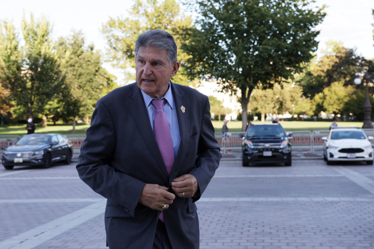 Sen. Joe Manchin, clutching the button of his suit jacket, stands near parked cars.