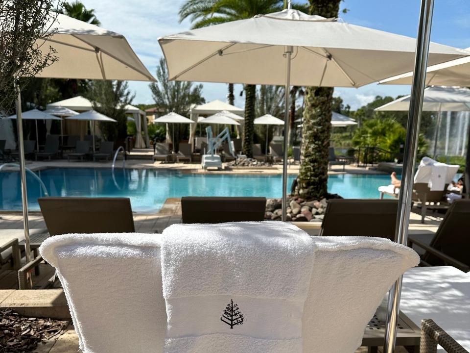 Photo from behind of a pool lounge chair with a white cover and towel resting on the back. In the background, the viewer can see a pool, brown lounge chairs, palm trees, and umbrellas.