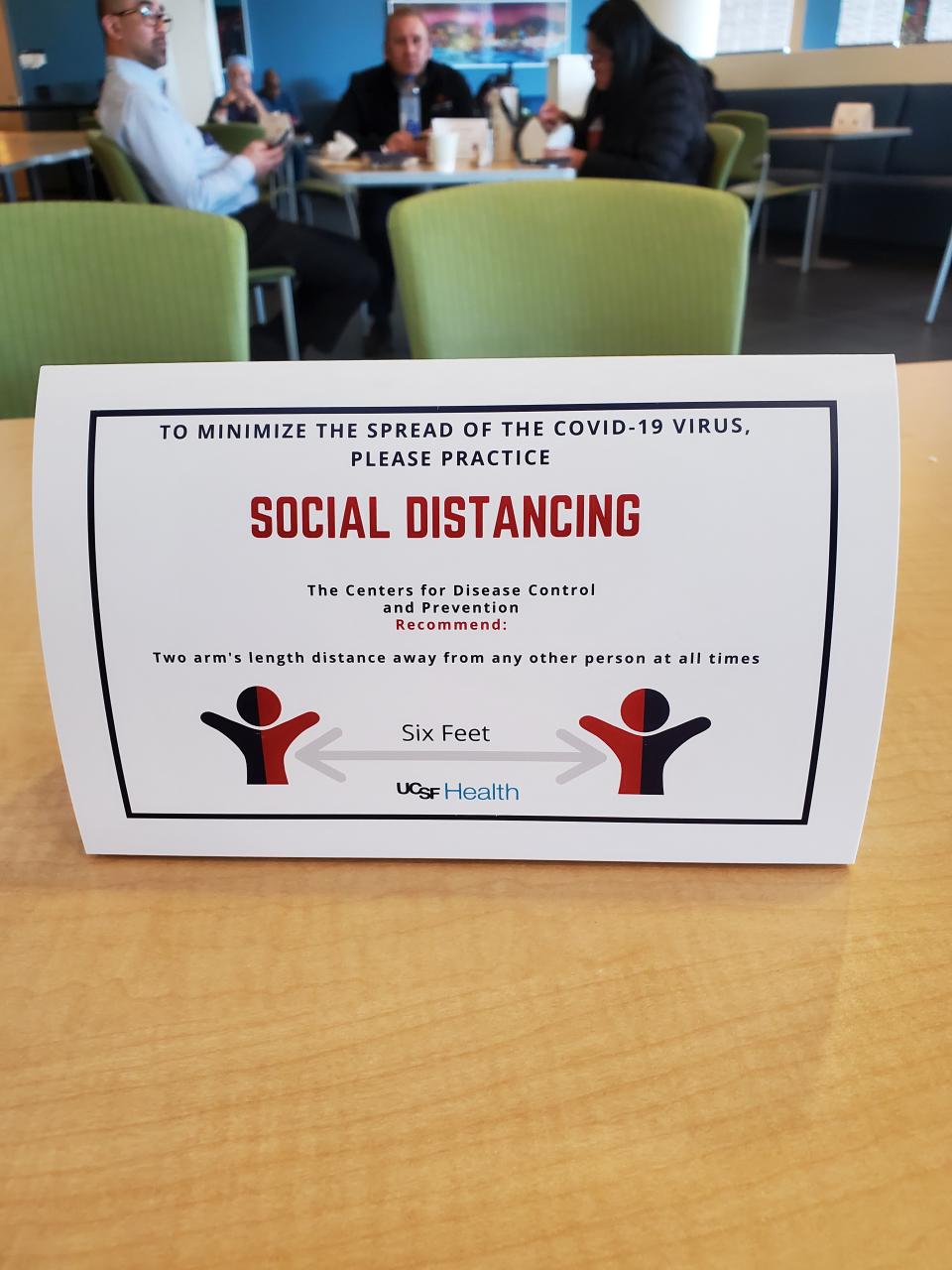 A sign on a table provides instructions for social distancing, while a group of people are seen in the background, selective focus, sitting around a small table, at a hospital in San Francisco during an outbreak of the COVID-19 coronavirus, California, March 16, 2020. (Photo by Smith Collection/Gado/Getty Images)