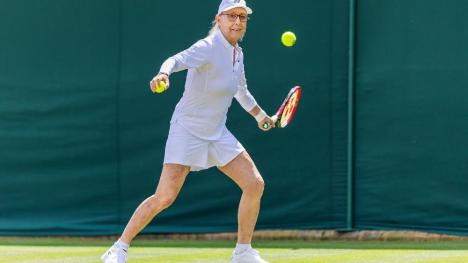 martina navratilova prepares to swing and hit a tennis ball with a red and black racket, she wears an all white outfit including a hat and shoes and holds a tennis ball in her other hand