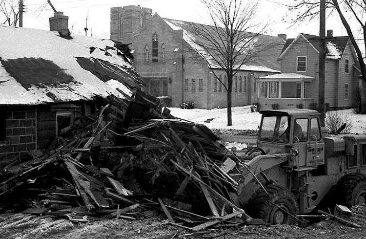 A bulldozer demolishes a home as part of the connector route clearance for I-496, January 1966.