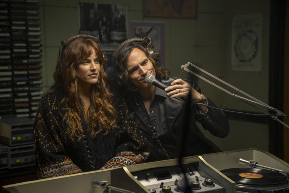 riley keough and sam claflin in character for daisy jones and the six sit in front of a microphone with headphones on