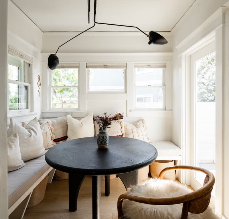 dining area with large cushioned bench with pillows, black round table, one chair with shag pillow, white walls, lots of windows