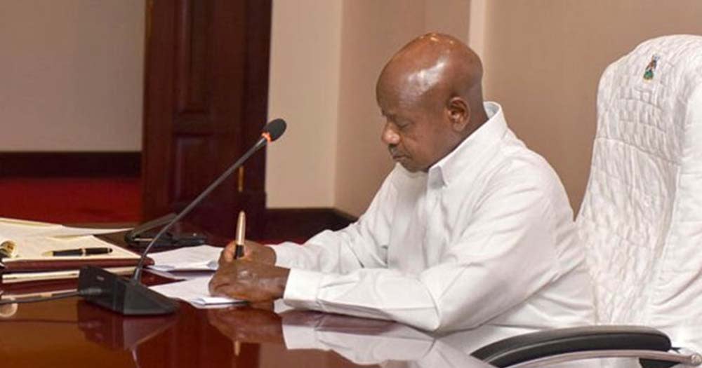 Ugandan President wearing white shirt sits at desk and signs anti-LGBTQ+ bill into law.