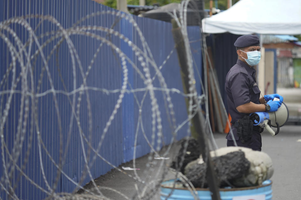 A police man stand next to coils of barbed wire near Top Glove factory hostel in Shah Alam, Malaysia, Wednesday, Nov. 25, 2020. Malaysia's Top Glove Corp., the world's largest maker of rubber gloves, says it expects delays in deliveries after more than 2,000 workers at its factories were infected by the coronavirus, raising the possibility of supply disruptions during the pandemic. (AP Photo/Vincent Thian)