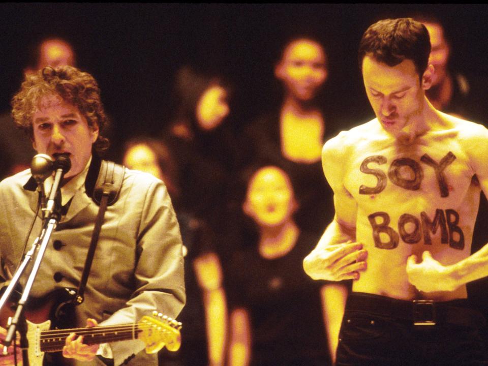 Bob Dylan singing and playing guitar while a man without a shirt on and the words "soy bomb" written on his chest in black paint is posing oddly behind him onstage.