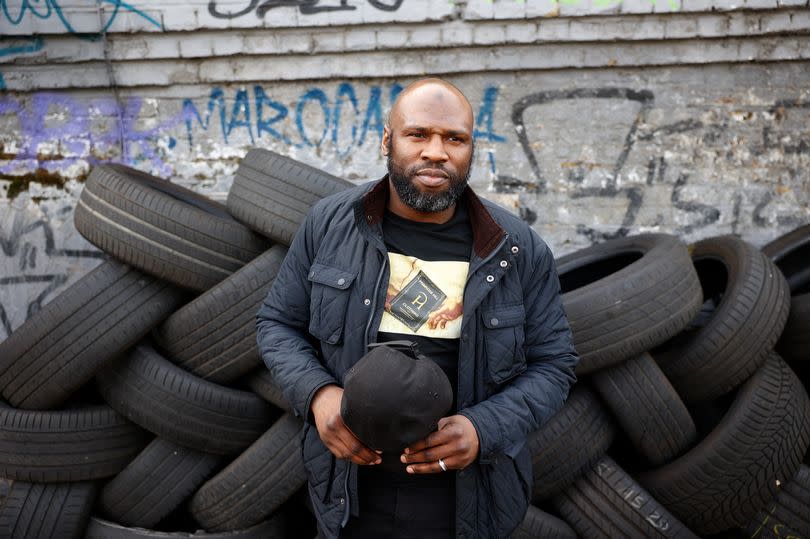 Dwaine Patterson in a jacket and holding his baseball cap in his hands in front of a pile of tyres