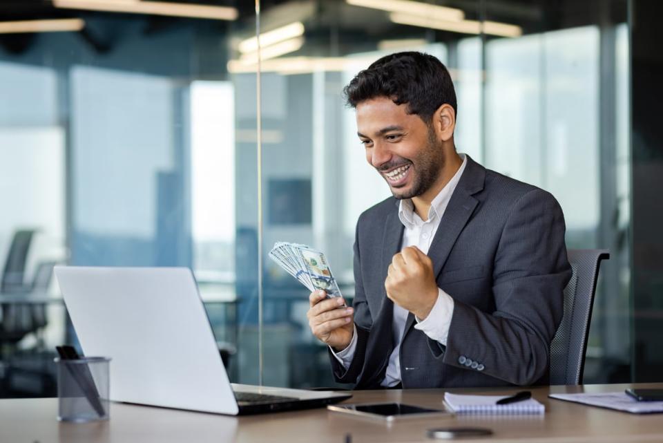 A smiling investor, holding money in one hand, looks at something on a laptop worth getting excited about.