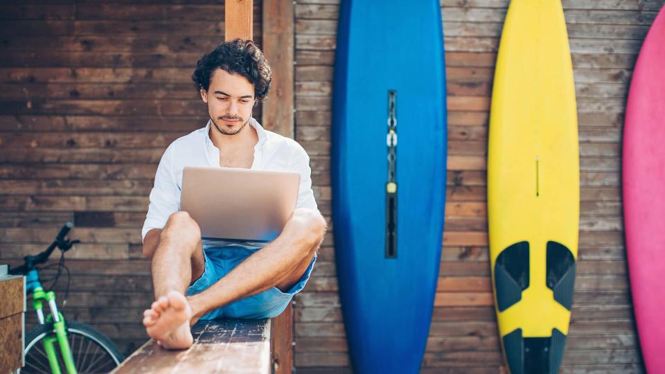 Young man using laptop outdoors in the summer with surfing boards in the background.