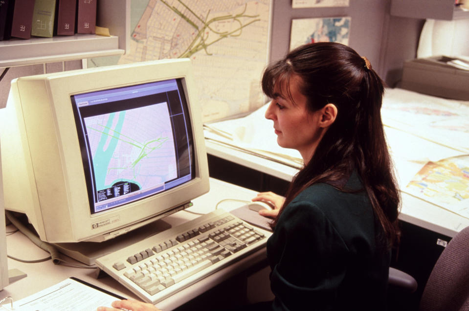 A person sits at a desk working on a computer that displays a map. The workspace is organized with maps and books around